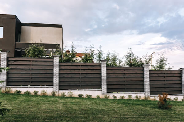 Horizontal tiered sections of brown wooden boards fence and white brick pillars. Live plantings. Green thuja bushes and lawn. Territory landscaping. Capital fencing. Hedge and wall on a country house.