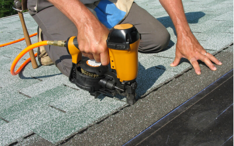 Carpenter uses nail gun to attach asphalt shingles to roof