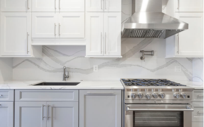 White kitchen built with shaker style cabinets and white quartz countertop. Shows stainless steel appliances, full granite backsplash. Polished Chrome faucet and hardware