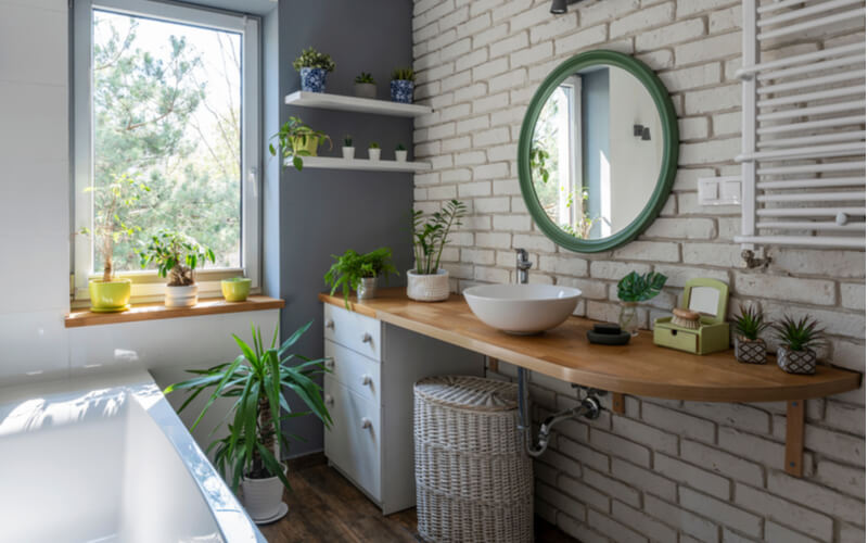 Industrial white bathroom with window, bath, brick wall, green plants and wooden counter. Loft interior of bathroom in apartment in scandinavian style.