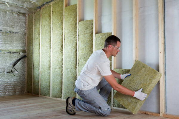 Worker in protective goggles and respirator insulating rock wool insulation in wooden frame for future house walls for cold barrier. Comfortable warm home, economy, construction and renovation concept