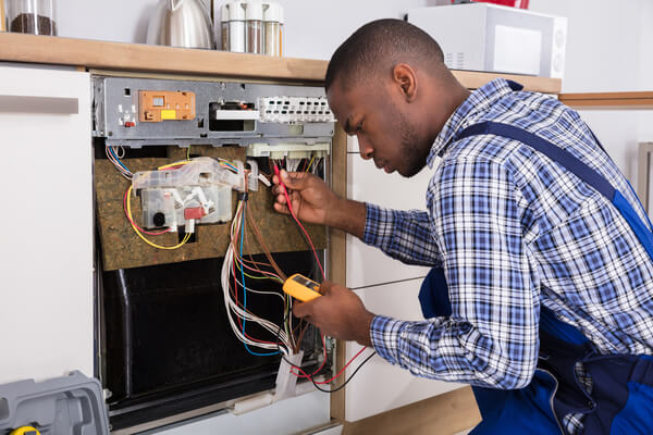 Young Male African Technician Fixing Dishwasher With Digital Multimeter In Kitchen