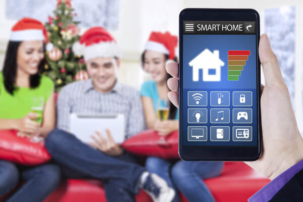 Hand holding a mobile phone with smart home system applications, shot with three young people wearing santa hat and using digital tablet in christmas day