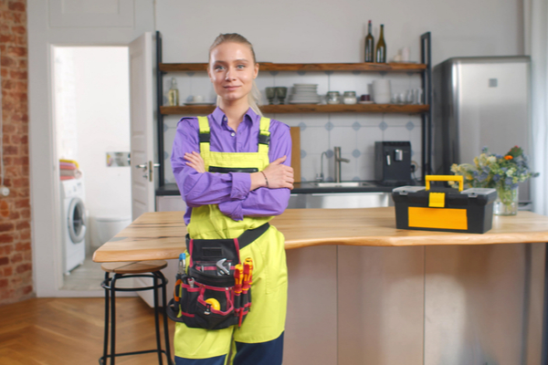 Portrait of beautiful young female handyman in overall and tool belt standing in home kitchen and smiling at camera. Repairman service and female hard work concept