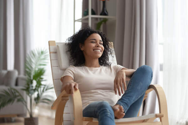 Smiling calm young black woman relaxing on comfortable wooden rocking chair in living room, happy healthy black girl enjoy breathing fresh air resting in armchair at home feel stress free on weekend
