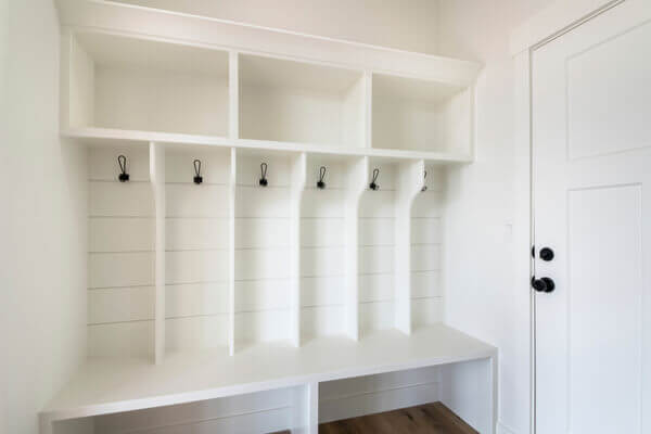 Interior of a white mudroom with white garage door and wooden flooring. Mudroom with white wooden bench and storage with black hanging hooks matching with the doorknob.